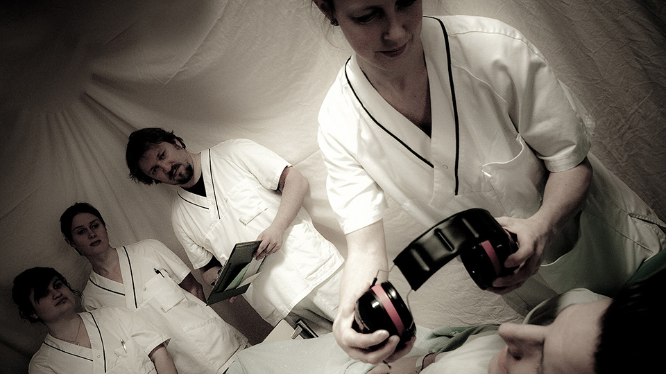Performers in white medical uniforms inside a draped clinic set, one holding a pair of black headphone-like devices over a reclining patient