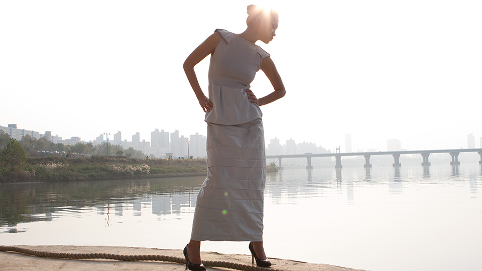 Model in a long pale grey dress with structured shoulders posing on a riverside walkway, Seoul skyline hazy in the background