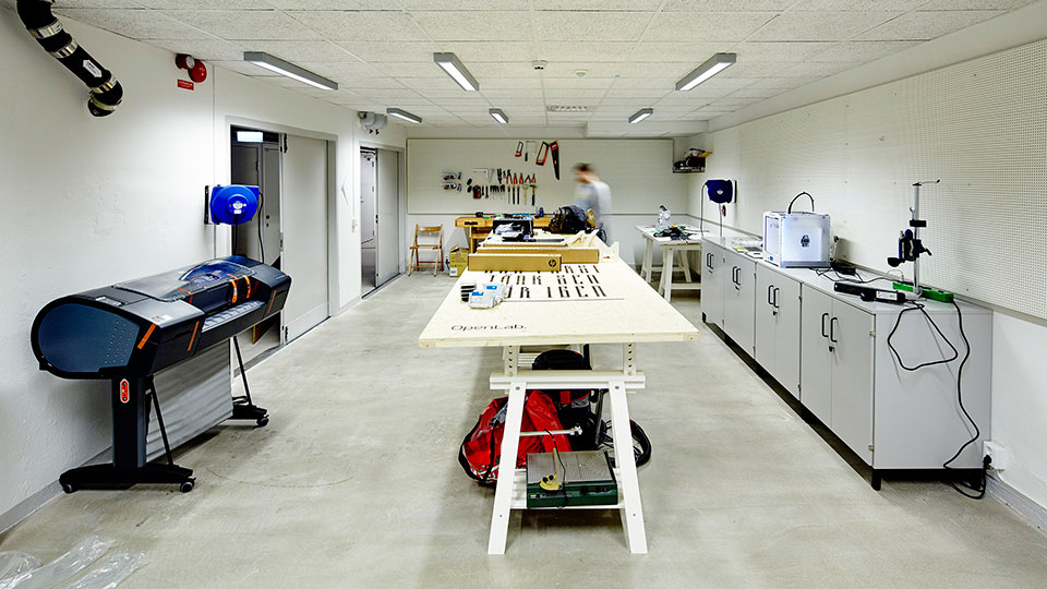 Bright workshop room with a long central worktable, large-format printer, 3D printer, and tools mounted on the back wall