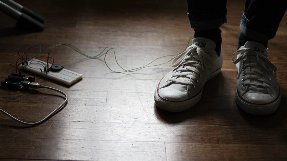 Close-up of white sneakers on a wooden floor next to a small electronics prototype with wires trailing across the floor