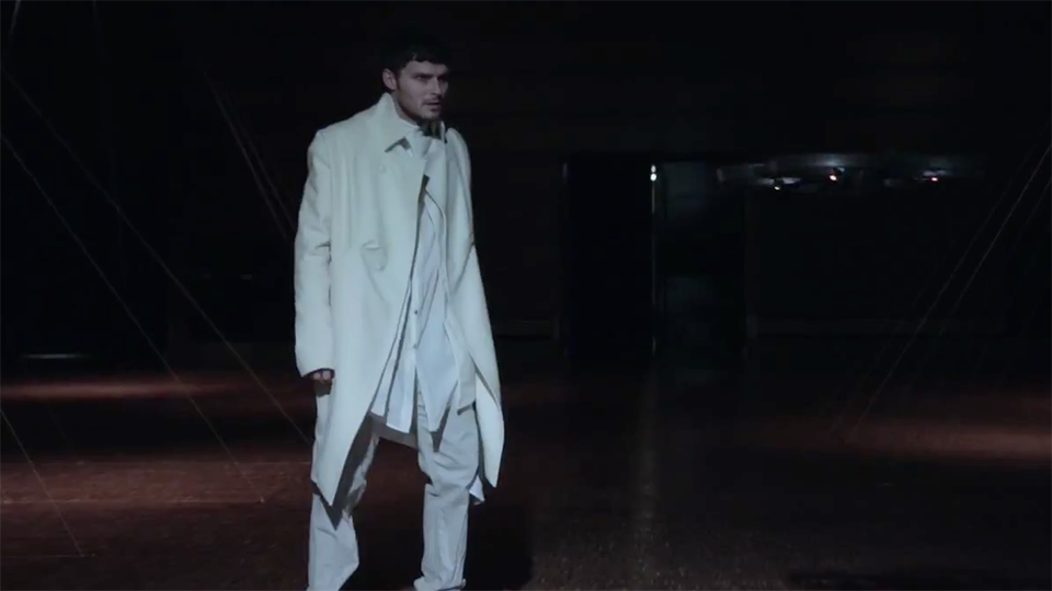 Video still: a model in a layered cream coat and trousers stands on a darkened runway lit from above