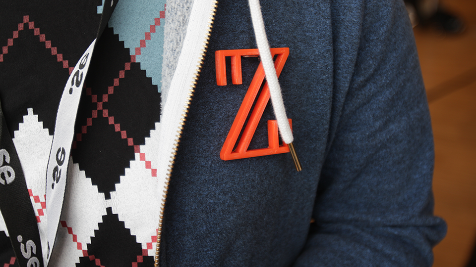 Close-up of an orange Ziggy Z-shaped lapel pin worn on a blue cardigan over an argyle shirt