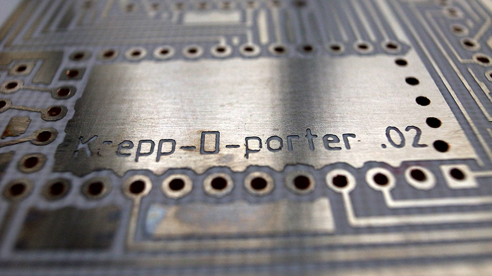 Macro shot of an etched circuit board engraved with the text 'Krepp-O-porter .02', showing copper traces and through-holes