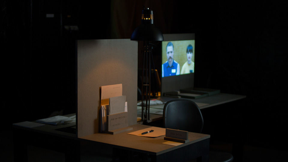 Dimly lit exhibition consultation booth with a desk lamp, document holder, name plate, and a video projection of two people in the background
