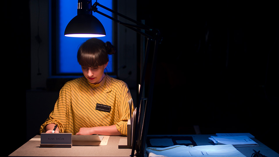 Woman in a yellow striped shirt with a name badge sits writing under a desk lamp, with a 'Nina Bačun' name plate in front of her
