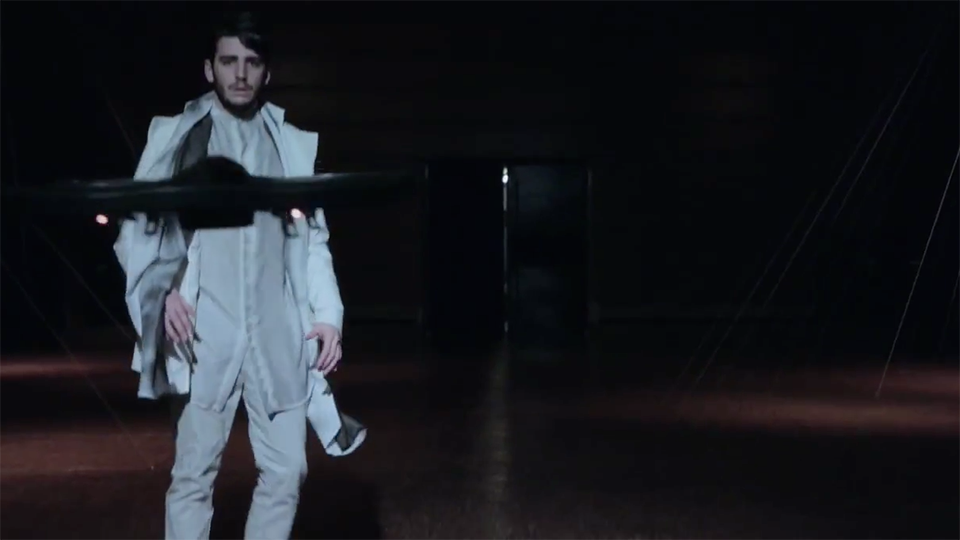 Video still: a model in a structured pale outfit walks forward as a small drone hovers in front of him