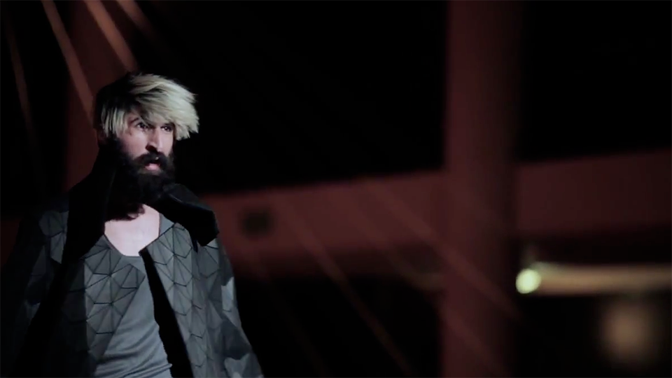 Video still: a bearded model with blonde hair wearing a geometric faceted grey top against a dark backdrop