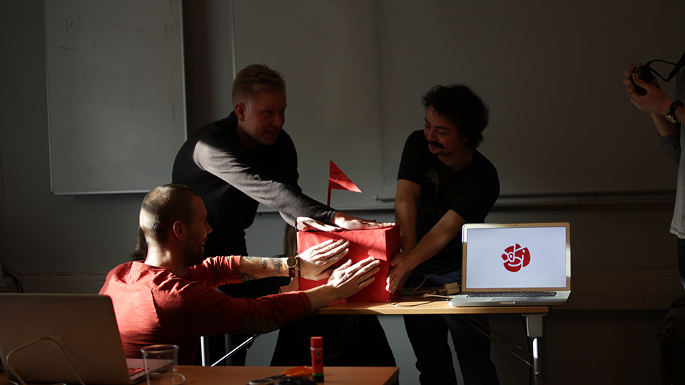 Three workshop participants gathered around a table examining a red and white prototype, with a laptop displaying a logo nearby