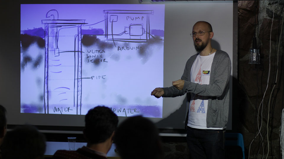 A bearded presenter at Stockholm Makerspace gesturing in front of a projected hand-drawn diagram of a water-level sensor