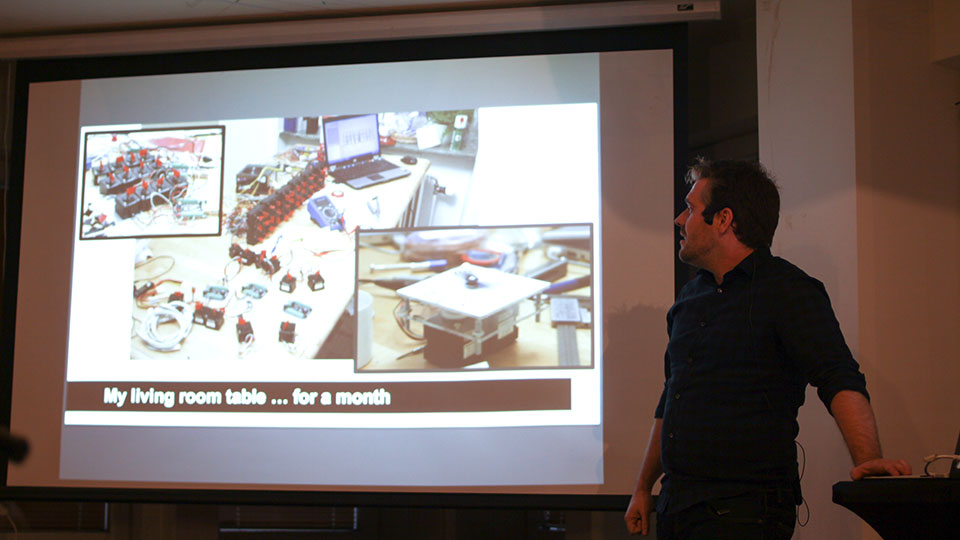 David Cox presenting beside a slide captioned 'My living room table … for a month' showing a workspace cluttered with prototypes