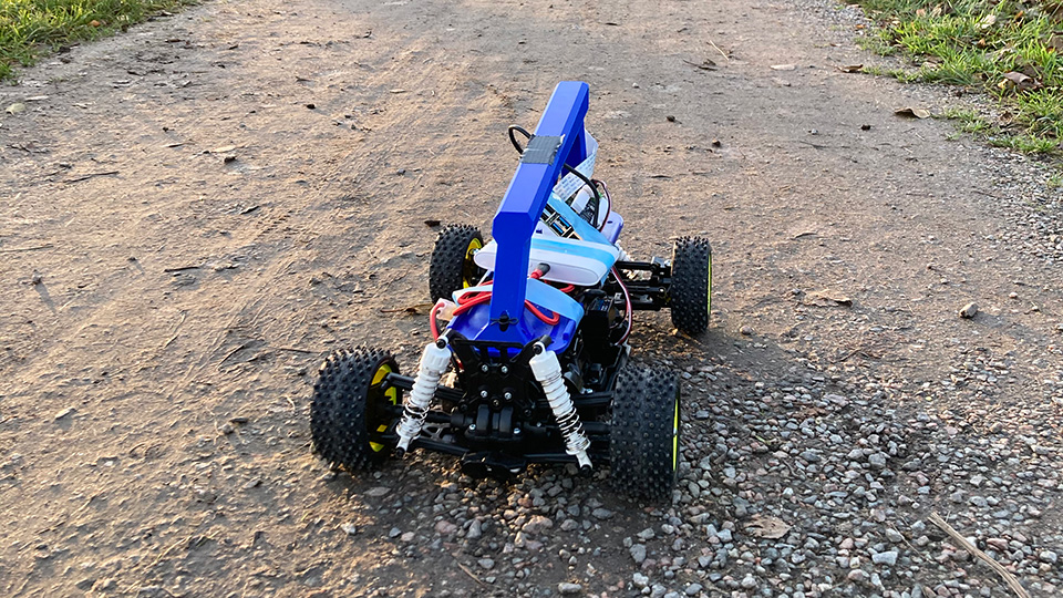 Blue 1/10 scale RC car fitted with a tall camera mast and electronics, parked on a gravel path