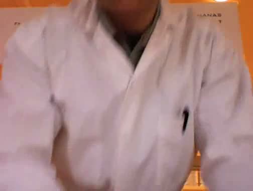 Video still: a person in a white lab coat seated in front of partially visible signage ending in 'nanas