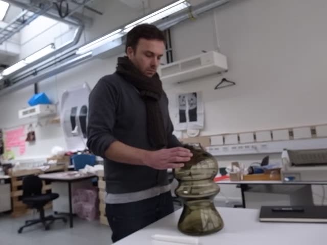 Video still: a man in a glass workshop holding a tall hand-blown green glass vessel on a worktable