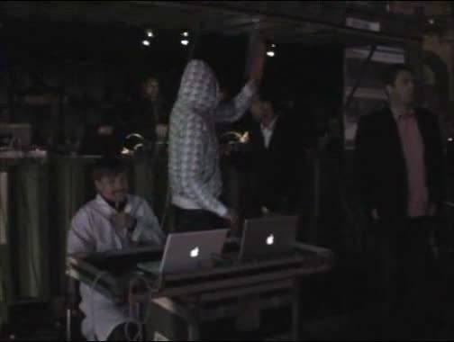 Video still: a hooded dancer with arm raised performs on stage behind a seated laptop performer, with onlookers in the background