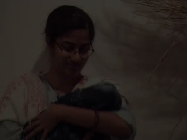 Video still: a woman in glasses and an embroidered white tunic cradling a dark bundle in a dimly lit room